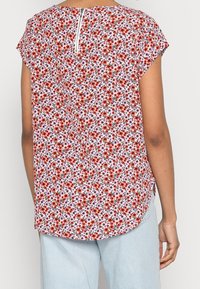 Blusa a maniche corte con stampa floreale in bianco, con fiori rossi e viola. Presenta una cerniera sul retro e un orlo arrotondato. Tessuto morbido e leggero.