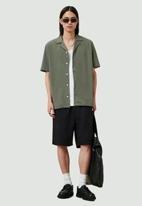 Jovem a usar óculos de sol, camisa de manga curta verde-azeitona sobre uma t-shirt branca, calções pretos, sapatos pretos com meias brancas, segurando uma mala preta.