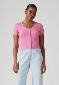 Gilet court à manches courtes rose côtelé avec un col en V et une rangée de boutons sur le devant, associé à un pantalon en denim bleu clair.