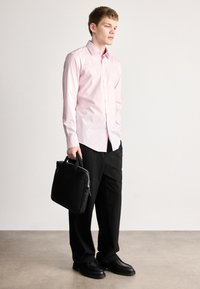 Φουξια πουκάμισο με κουμπιά, μαύρο παντελόνι και μαύρα παπούτσια. Κρατώντας μια μαύρη, δομημένη τσάντα με λαβές στην κορυφή. Απλός, ραμμένος σχεδιασμός.