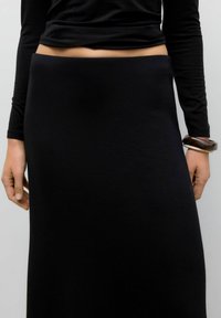 Persona che indossa una maglietta nera a maniche lunghe e una gonna nera a vita alta, con due bracciali spessi sul polso destro contro uno sfondo semplice.