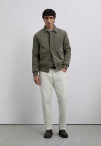 Chaqueta de pana verde oliva con bolsillos frontales, combinada con chinos beige claro y zapatos oscuros de tipo slip-on, de pie contra un fondo neutral.