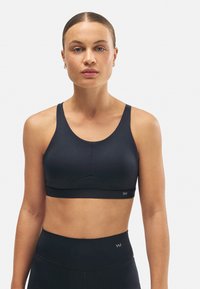 Sujetador deportivo negro hecho de tela suave, con una banda ancha, diseño de espalda nadadora y costura central sutil. Logo visible en la banda.