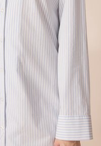 Camicia a righe blu chiaro e bianca con bottone frontale e maniche lunghe. Il tessuto è liscio e il polsino presenta un motivo a righe.