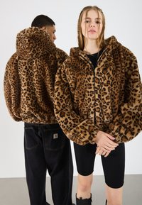 Două persoane îmbrăcate în jachete cu imprimeu de leopard maro din blană artificială, una cu fața spre cameră și cealaltă cu spatele întors, într-un spațiu interior neutru.