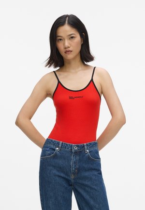 Femme aux cheveux sombres mi-longs, portant un haut rouge à fines bretelles avec une bordure noire et un jean bleu taille haute, debout les mains dans le dos.