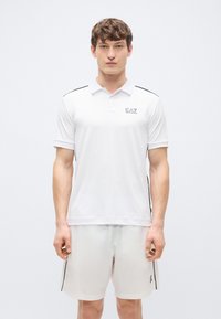 Weißes Poloshirt mit kurzen Ärmeln, schwarzen Akzentstreifen und EA7-Logo auf der Brust. Kombiniert mit passenden weißen Shorts.