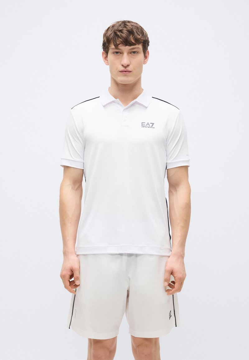 Weißes Poloshirt mit kurzen Ärmeln, schwarzen Akzentstreifen und EA7-Logo auf der Brust. Kombiniert mit passenden weißen Shorts.