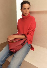 Red turtleneck sweater with ribbed texture paired with denim jeans. A matching red handbag with a gold accent rests on the lap.