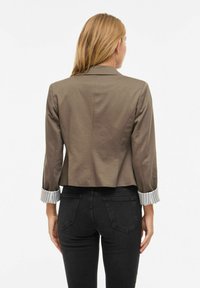 Femme blonde portant un blazer olive court avec des manches rayées retroussées et un jean noir, vue de dos sur un fond blanc.