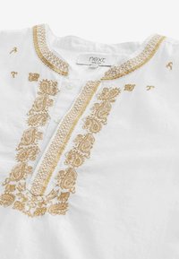 Camicia bianca in lino con ricami oro a motivo paisley attorno al colletto e alla listino dei bottoni, con due bottoni bianchi e un'etichetta "next" all'interno del colletto.