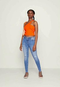 Top cropped laranja com um ombro só e detalhe estampado; combinado com jeans skinny desfiados de tom azul claro. Usado com sandálias rasas sobre um fundo neutro.