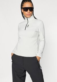 Top blanco de manga larga con cremallera y cuello alto, acompañado de pantalones negros con cremalleras laterales y un acabado texturizado.