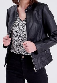 Veste en simili cuir noir avec un col rond, une fermeture éclair argentée et des poches latérales, portée sur un haut floral noir et blanc.