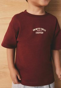 Bordo bombažna majica s kratkimi rokavi in okroglim izrezom, z kremno obarvanim potiskom z napisom "NEOMEJENI NASMEHI VSAK DAN."