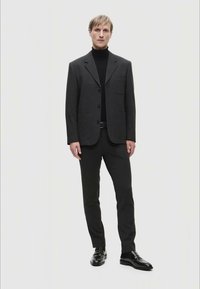 Giacca da abito grigia scura con rever a punta e tasca anteriore, abbinata a un dolcevita nero e pantaloni coordinati, completata da scarpe nere lucide.