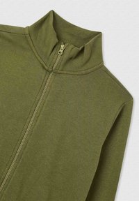 Giacca con zip color verde oliva, collo alto, texture a coste e chiusura frontale con zip. Tessuto liscio con vestibilità rilassata.