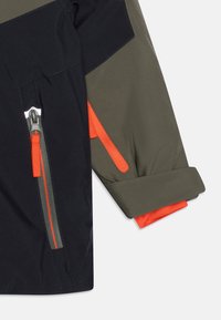 Nepremokavá bunda v čiernej a olivovo zelenej farbe, so zipsovými vreckami s oranžovými akcentmi a srolovanými rukávmi.