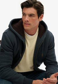 Man in een donkerblauwe ritsjack met een bruin fleece voering over een crèmekleurige trui, zittend en kijkend opzij tegen een effen achtergrond.