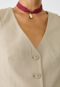 Beżowy, w paseczki, bezrękawnik z dekoltem w kształcie litery V, z dwiema okrągłymi guzikami, w połączeniu z magentową koronkową chokerą ozdobioną złotym wisiorkiem.