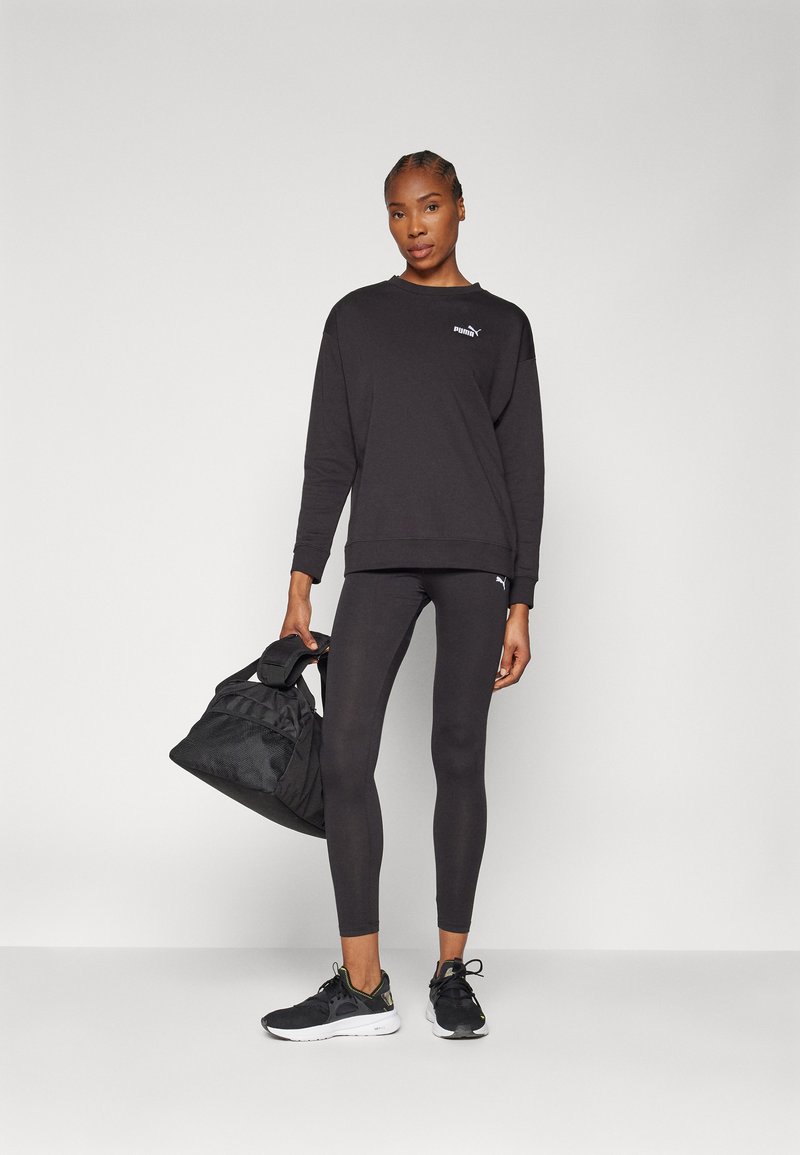 Schwarzes Sweatshirt mit Logo, schwarze Leggings und schwarze Sneakers. Das Modell hält eine schwarze Sporttasche. Einfaches, sportliches Design mit einer figurschmeichelnden Form.