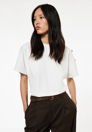 Frau mit langem dunklem Haar, die eine weiße kurzärmelige Bluse und hoch taillierte dunkelbraune Hose trägt, mit den Händen in den Taschen.