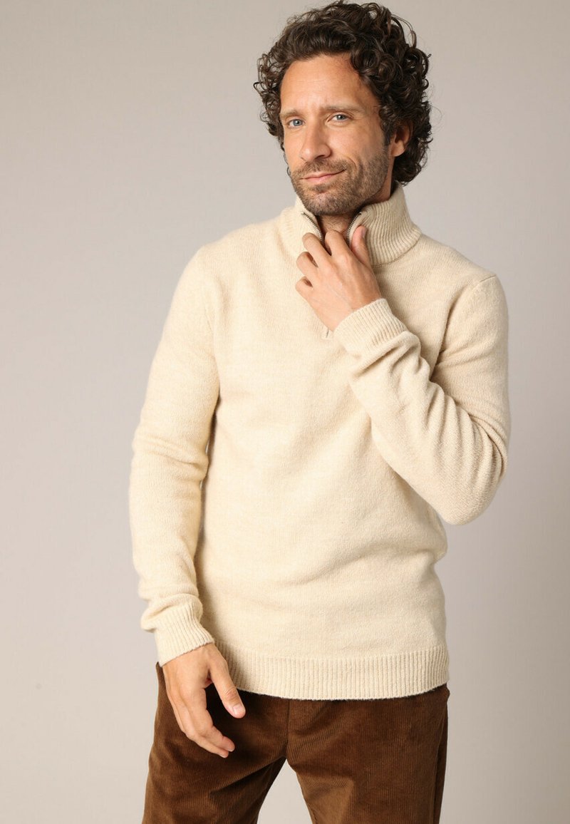 Beige coltrui gemaakt van zacht materiaal met geribbelde manchetten en zoom, gecombineerd met een bruine corduroy broek.
