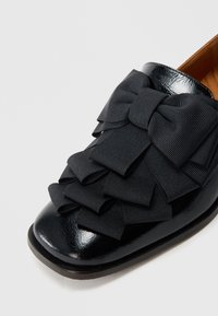 Chaussure en cuir verni noir dotée d'une décoration composée de nœuds en ruban noir superposés à l'avant. Bout arrondi et talon bas.