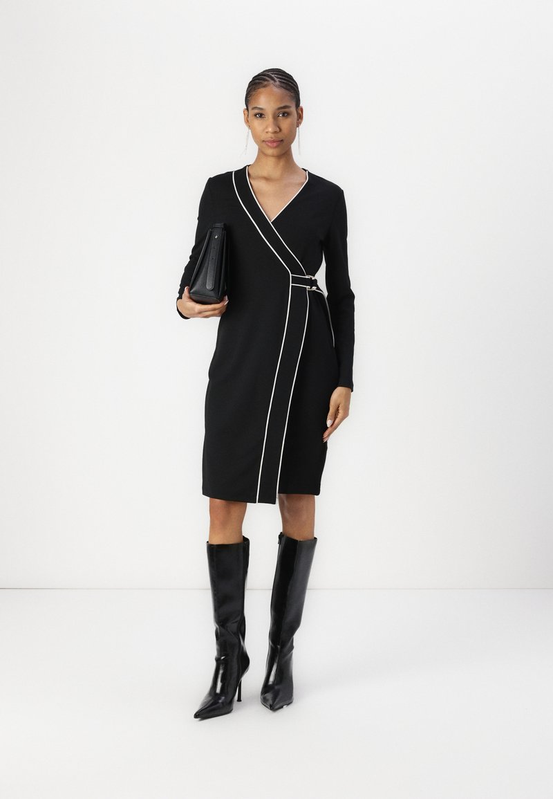 Vestido negro de manga larga con cierre cruzado y detalles de ribete blanco, de longitud a la rodilla, combinado con botas altas negras de brillo y un bolso estructurado negro.