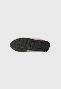 Semelle extérieure en caoutchouc noir crantée avec des motifs carrés et rectangulaires, fixée à une base de chaussure beige avec une finition texturée et un design minimal.