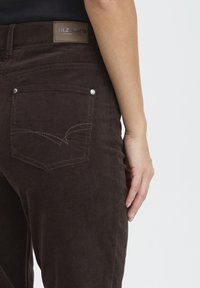 Brown corduroy pants with a fitted design, featuring a logo patch, silver hardware, and back pocket detailing. Textured fabric.