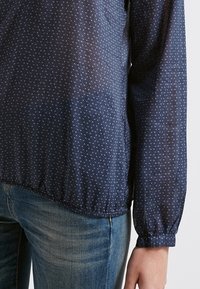 Náměsíčně modrá blůza z lehké texturované tkaniny, která má malý trojúhelníkový vzor a elastické manžety na rukávech.