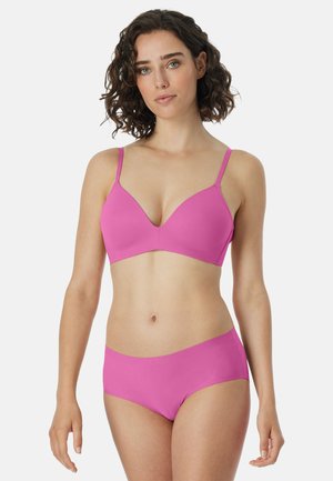 Woman with curly brown hair wearing a plain pink bra and matching seamless panties, standing against a white background.