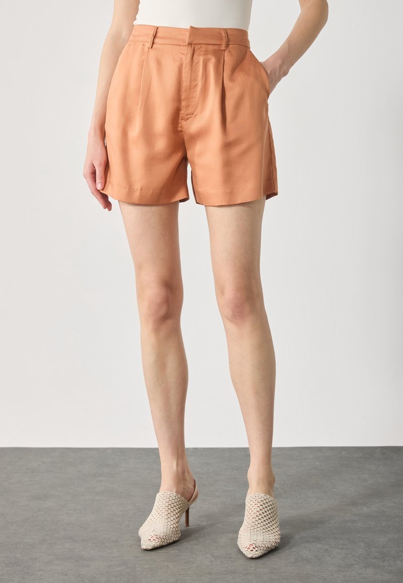 Femme portant un short orange plissé taille haute avec poches latérales et des mules blanches tissées à talons hauts, debout sur un sol gris.