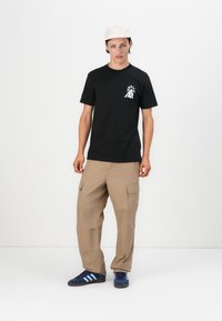 Zwarte T-shirt met een witte graphic op de linkerkant van de borst, khaki cargo shorts en marineblauwe sneakers met lichtblauwe strepen. Beige pet.