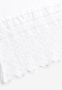 Witte katoenen blouse met ingewikkelde opengewerkte kanten zoom, bloemmotief en geborduurd logo. Zachte textuur en detail met gegolfde rand.