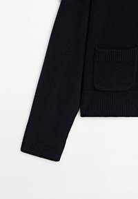 Manche et partie inférieure d'un pull noir en maille avec poignets côtelés et poche avant sur fond blanc.