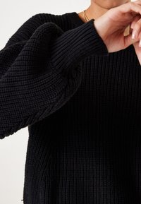 Schwarzer gestrickter Pullover mit einer dicke Rippstruktur, weit geschnittenen Ärmeln und einem klassischen Rundhalsausschnitt. Nahaufnahme, die Handbewegungen zeigt.