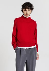 Maglione rosso a collo alto, corto, realizzato in un materiale a maglia liscia, indossato sopra una camicia bianca con colletto, abbinato a pantaloni grigi a vita alta con pieghe.