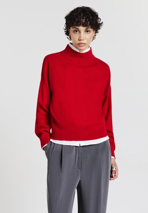 Maglione rosso a collo alto, corto, realizzato in un materiale a maglia liscia, indossato sopra una camicia bianca con colletto, abbinato a pantaloni grigi a vita alta con pieghe.