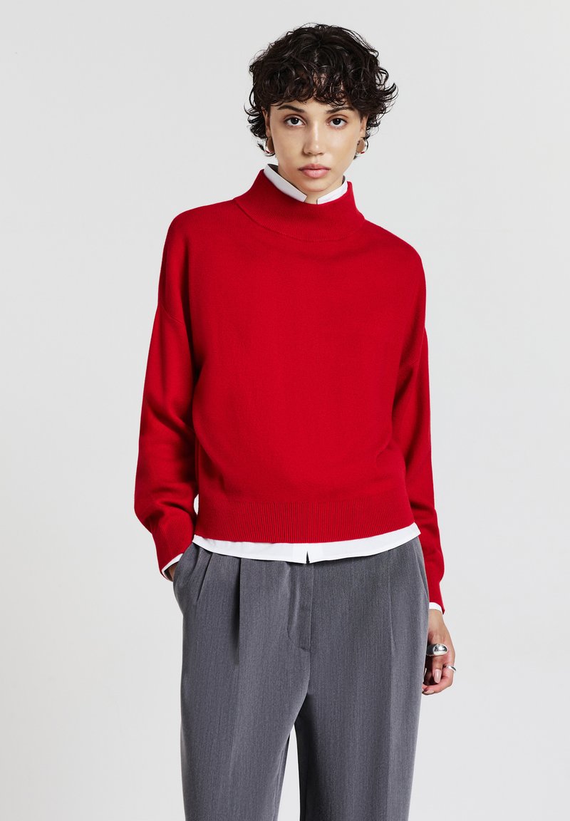 Maglione rosso a collo alto, corto, realizzato in un materiale a maglia liscia, indossato sopra una camicia bianca con colletto, abbinato a pantaloni grigi a vita alta con pieghe.