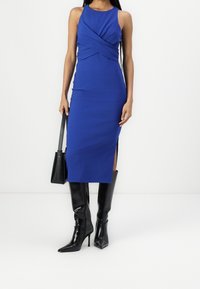 Kungablå, kroppsnära klänning med en tvistad framdesign, sidoblistar, i kombination med sleek svarta knähöga stövlar med klack och en liten svart väska.