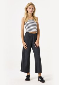 Zwarte geplooide brede broek met een elastische tailleband, gecombineerd met een wit en zwart gestreept crop top. Model draagt zwarte sportschoenen.