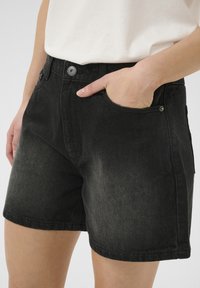 Crne traper kratke hlače s uskim krojem, koje imaju prednje džepove i metalne detalje. Tkanina ima blago izbjeljenje i teksturu.