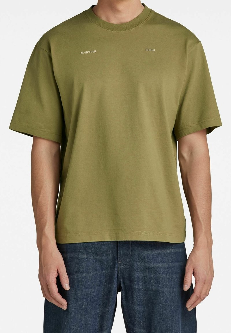 T-shirt manches courtes vert olive avec les inscriptions « G-STAR » et « RAW » imprimées près du col, porté avec un jean bleu foncé.