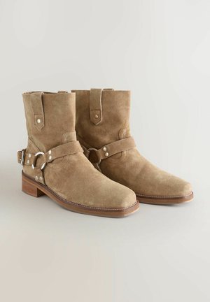 Stivale in pelle scamosciata color nocciola con punta arrotondata, dotato di una fascetta e un dettaglio metallico, e un tacco in legno basso e impilato.