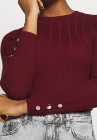 Pull côtelé bordeaux avec manches longues, présentant des accents métalliques et des détails de boutons aux poignets. Texture douce, coupe ajustée.