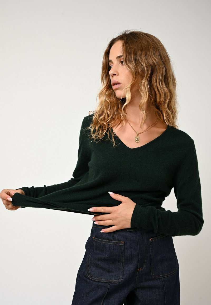 Dunkelgrüner V-Ausschnitt-Pullover aus weichem Stoff, mit langen Ärmeln und lässigem Schnitt, kombiniert mit dunklen Jeans.
