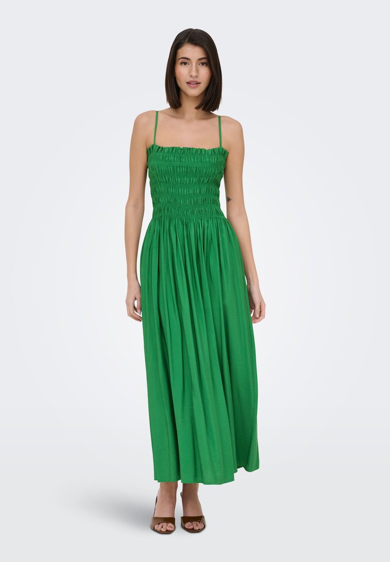 Femme aux cheveux foncés mi-longs portant une robe maxi verte sans manches avec un corsage froncé et des talons ouverts marron, debout sur fond blanc.