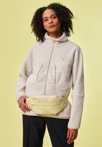 Cremfarbene Fleecejacke mit hohem Kragen, zwei Brusttaschen und Reißverschluss, kombiniert mit einer gelben Bauchtasche mit Reißverschluss und Akzenten.